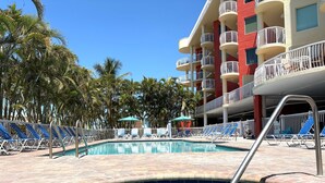 A swimming pool with lounge chairs and umbrellas, surrounded by palm trees and a multi-story building with balconies. - Last-Minute August Beach Deals – Save Big! (Treasure Island)