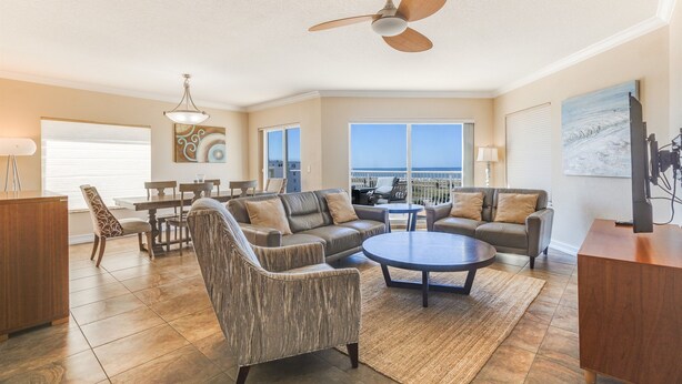 A spacious living room with a large sofa, a coffee table, and a dining area with chairs and a table. There is a ceiling fan, a TV, and a painting on the wall. - Last-Minute August Beach Deals – Save Big! (Treasure Island)