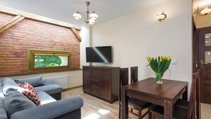 A living room with a brick wall, a flat-screen TV, a dining table with chairs, a sofa, and a vase with flowers.