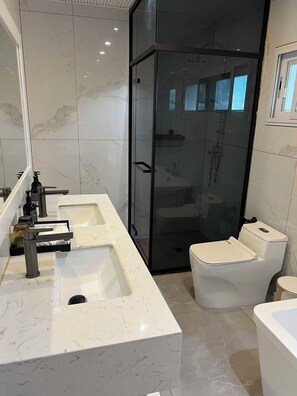 Um banheiro moderno com um espelho grande, duas pias, um chuveiro com box de vidro e um vaso sanitário.
