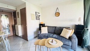 A living room with a grey sofa, two small round tables, a door, and a wall-mounted clock. - Argelès-sur-Mer - T2 Apartment with Balcony and Parking (Argelès-sur-Mer)