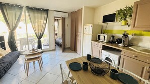 A compact living space with a dining area, kitchenette, and a balcony with curtains. - Argelès-sur-Mer - T2 Apartment with Balcony and Parking (Argelès-sur-Mer)