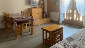 A cozy room with a wooden dining table, chairs, a small wooden table, a TV on a wooden stand, a radiator, and a window with curtains. - Functional studio for 4, West facing in Valloire (Valloire)