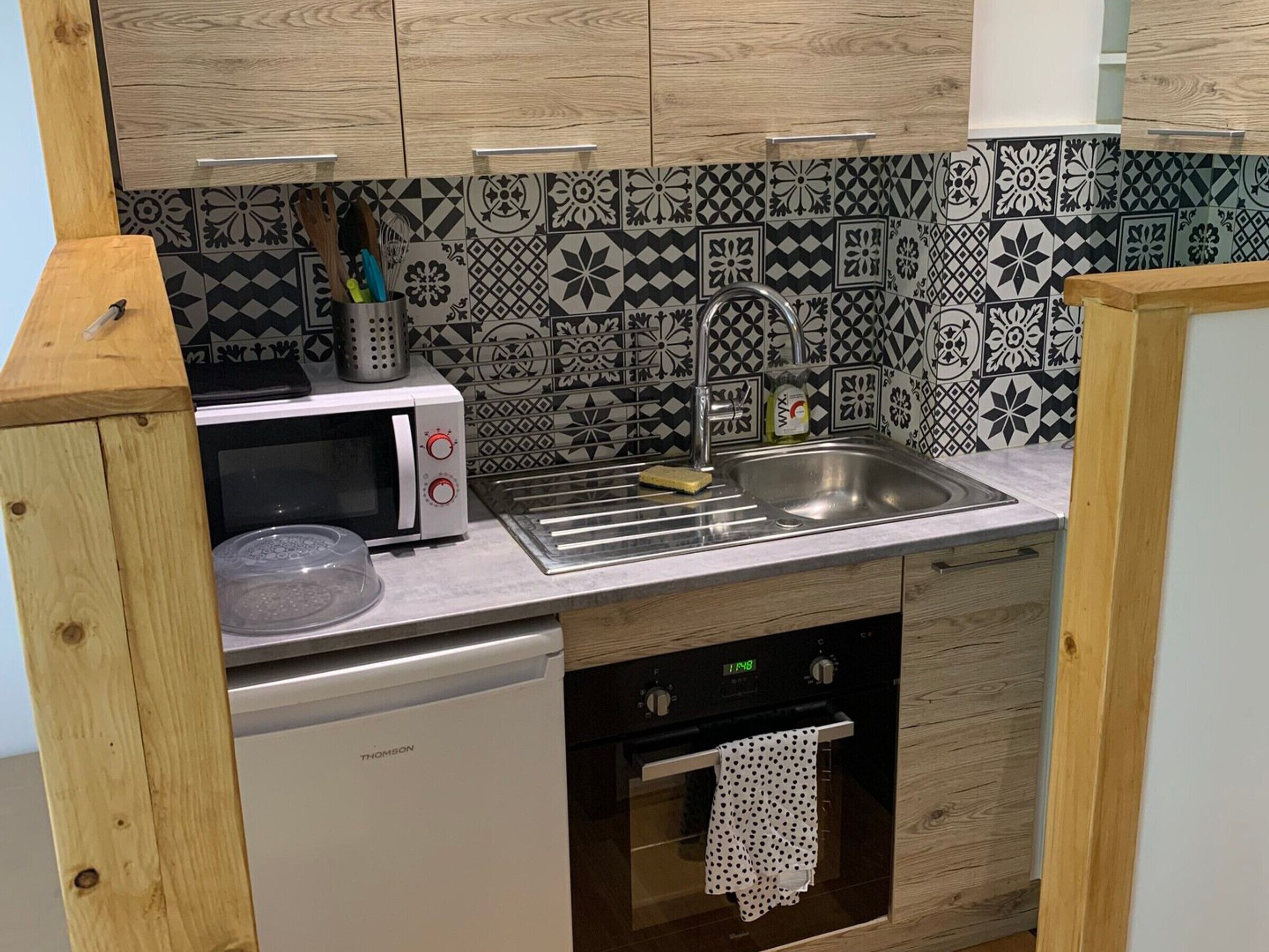 A compact kitchen with a microwave, sink, and dishwasher. The backsplash features a black and white geometric tile pattern.
