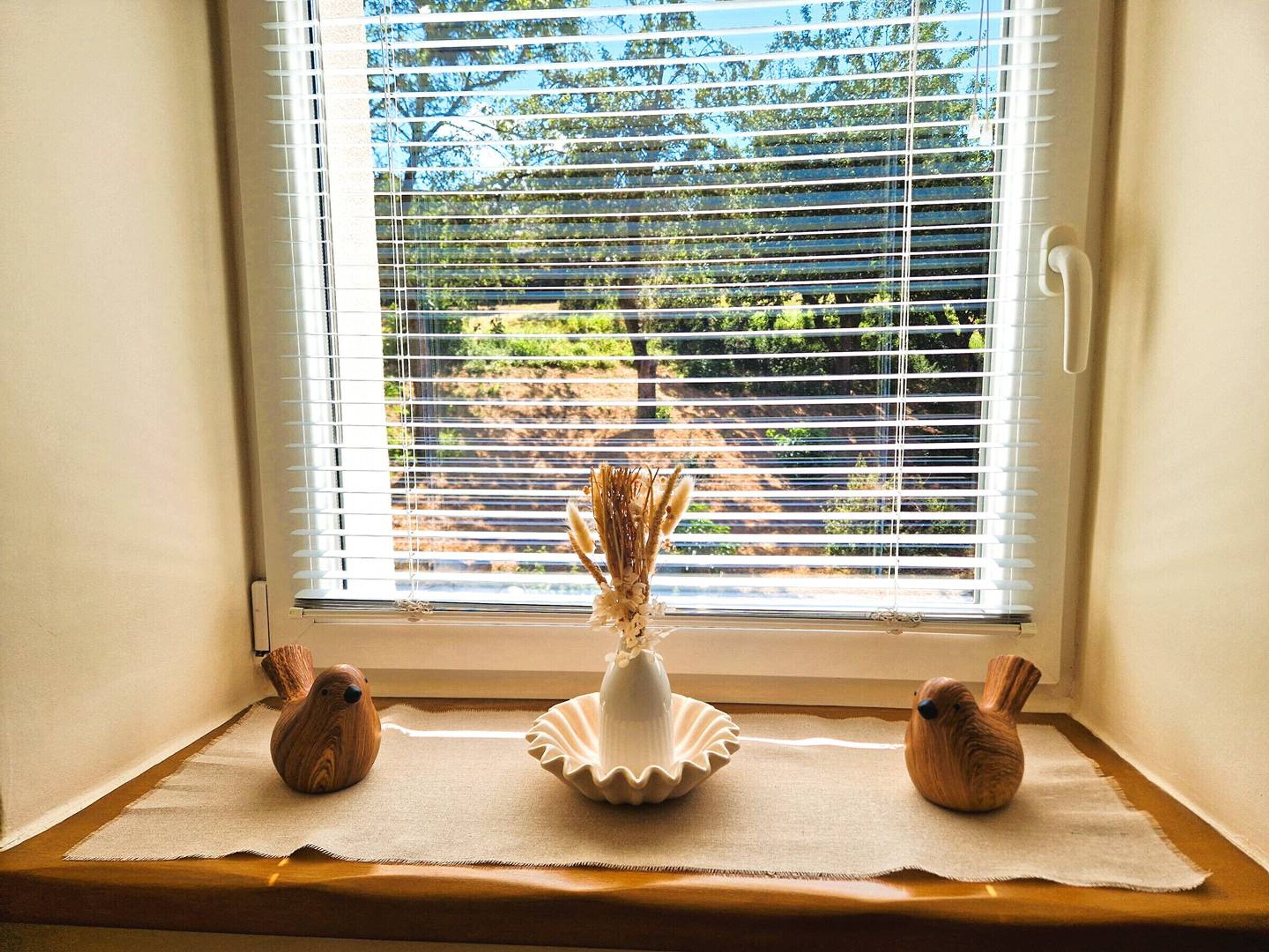 Une fenêtre avec des stores blancs, une étagère en bois ornée de trois vases en forme d’oiseaux et un vase blanc contenant des plantes séchées.