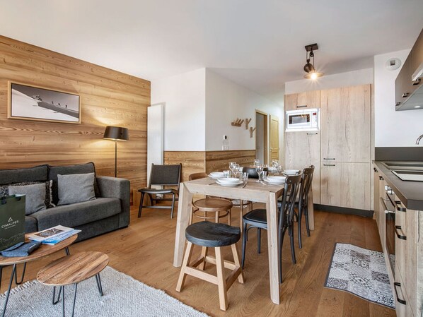 A modern living room with a sofa, a dining table set for four, and a kitchen area with wooden cabinets and a microwave. - 2BR Apartment in Les Saisies, sleeps 6 (Les Saisies)