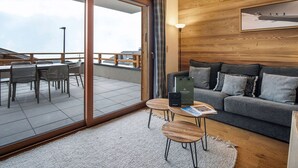 A modern living room with a grey sofa, wooden coffee table, and a glass door leading to a balcony with outdoor seating. - 2BR Apartment in Les Saisies, sleeps 6 (Les Saisies)