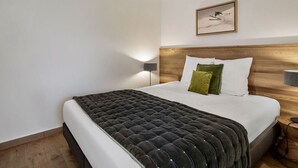 A modern bedroom with a wooden headboard, a large bed with a dark quilt, white and green pillows, and a wall-mounted light. - 2BR Apartment in Les Saisies, sleeps 6 (Les Saisies)