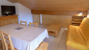 A room with a wooden ceiling, a television on a cabinet, a table with chairs, and a yellow armchair.