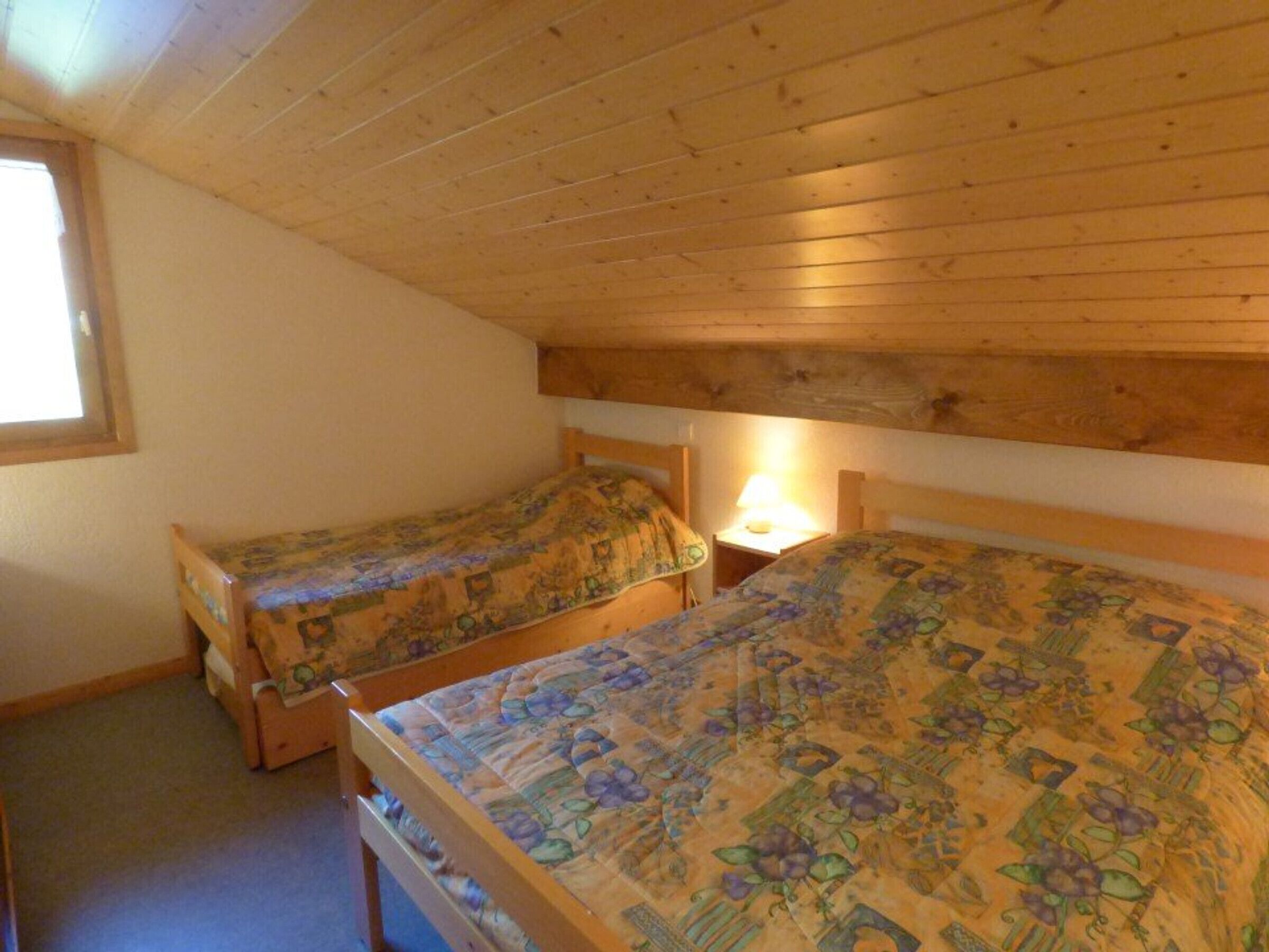 Une petite pièce chaleureuse, avec deux lits en bois, chacun agrément é de draps à motifs floraux. Une fenêtre se trouve sur le mur de gauche, et une table de chevet avec une lampe est placée entre les deux lits.