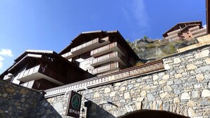 Un bâtiment en pierre doté de nombreux balcons, une enseigne indiquant un centre de sports d’hiver, et un ciel bleu dégagé.
