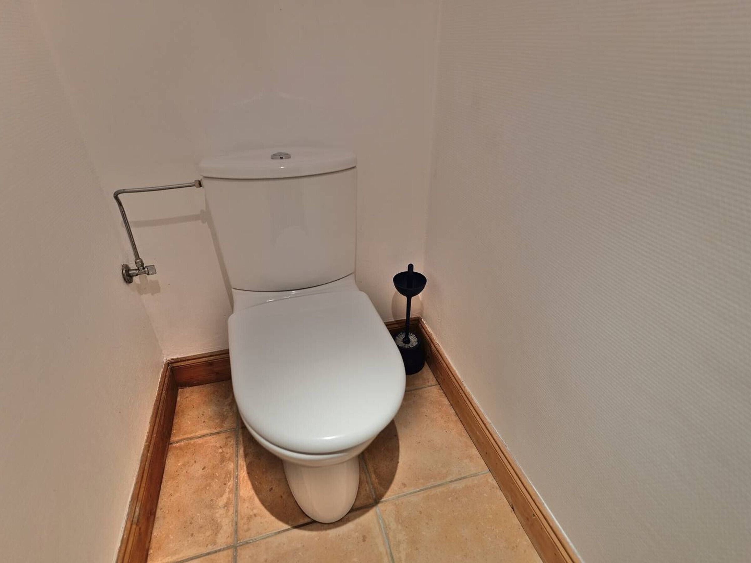 A small, white toilet with a tank and seat, positioned in a corner with a tiled floor and a wooden baseboard.