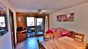 Un coin repas avec une table et des chaises en bois, un téléviseur et une porte-fenêtre coulissante donnant sur une terrasse extérieure.