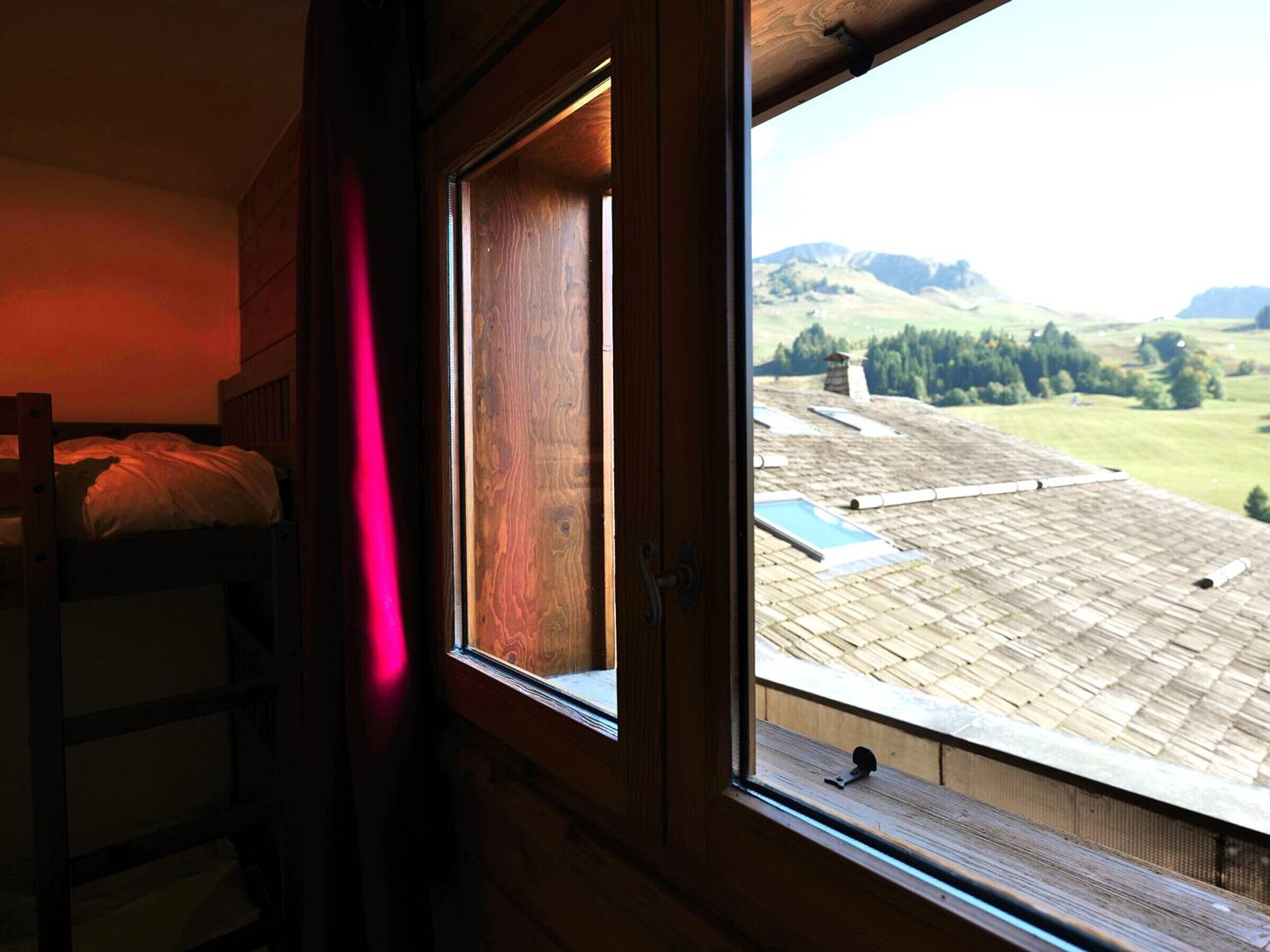 A room with a wooden bunk bed, a window with a view of a hilly landscape, and a red curtain.