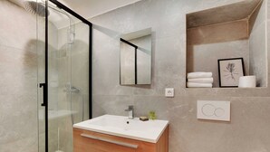 A modern bathroom with a glass shower enclosure, a wooden vanity with a white sink, and a shelf with towels and a framed botanical print. - Elegant Apartment - 2bd/6p - Hotel de Ville (Paris)