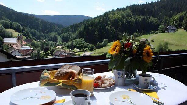 A table with breakfast items, a vase of sunflowers, and a scenic view of a village and mountains. - Apartment 'Margot Armbruster' with Mountain View, Balcony and Wi-Fi (Bad Rippoldsau-Schapbach)