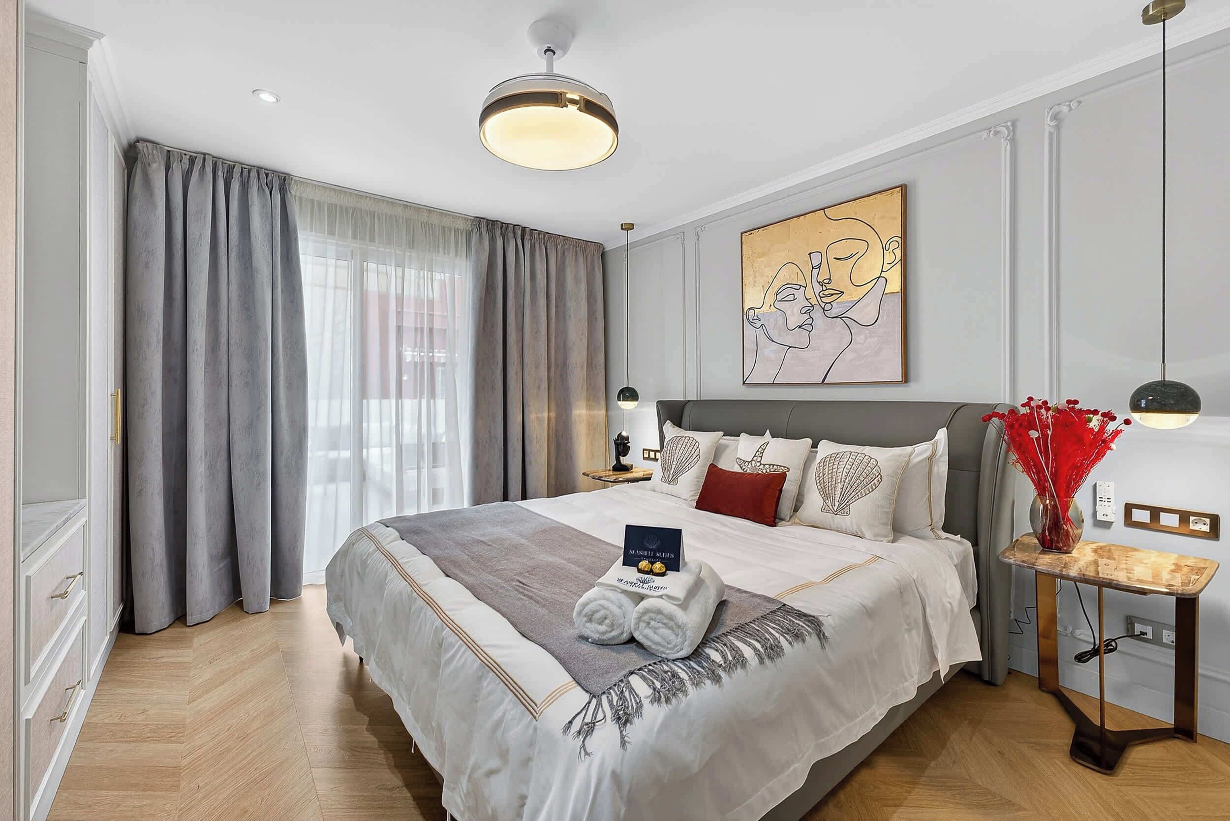 Hotellihuone, jossa on suuri sänky, yöpöytä lampun kanssa, taulu seinällä ja ikkuna, jossa on verhot.