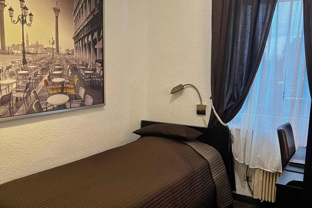 A hotel room with a bed, a desk, a chair, a lamp, a window with curtains, and a cityscape painting on the wall.