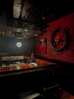 Un bar con mostrador de madera, estantes con botellas, una rueda de barco y un cartel en la pared.