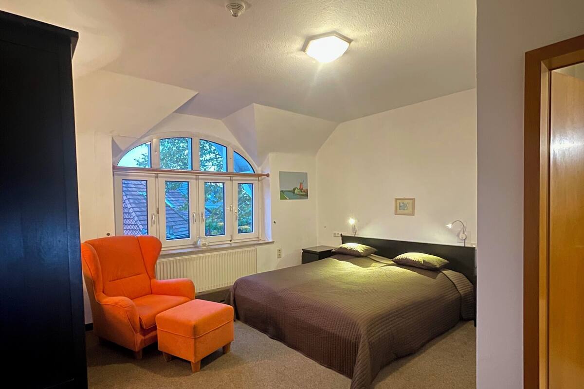 A bedroom with a bed, an orange chair, a window with a view of trees, and a ceiling light.