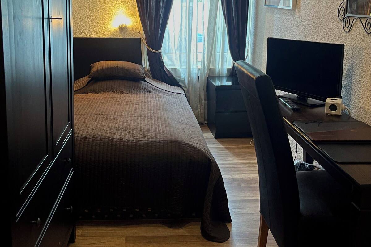 A bedroom with a bed, a desk, a chair, a TV, and a window with curtains.