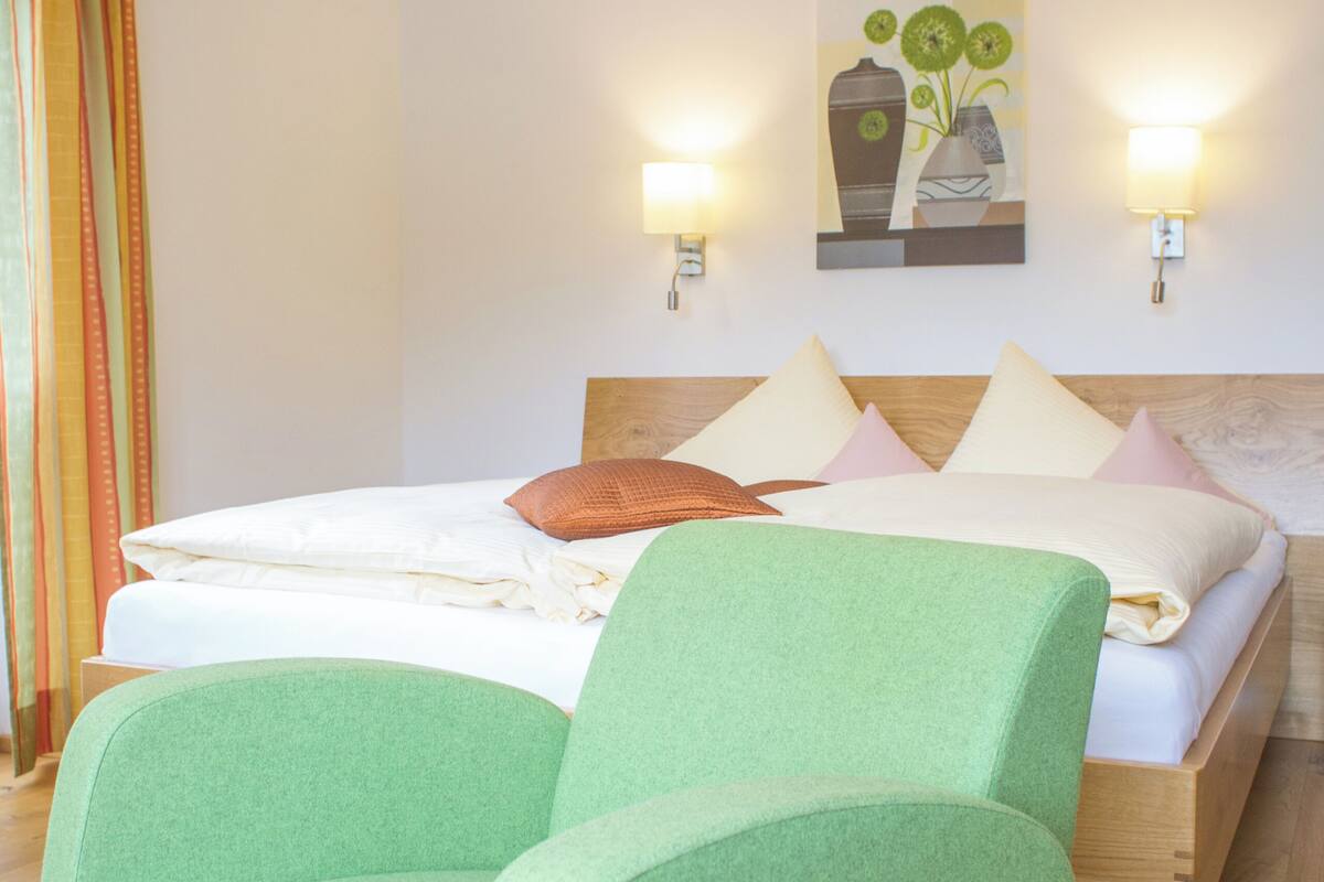Habitación de hotel con una cama, dos lámparas adosadas a la pared, un cuadro y un sillón verde.
