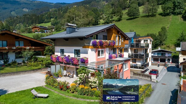 Une maison moderne à deux étages, dotée d’un balcon agrémenté de fleurs aux couleurs vives, d’une véranda en verre et offrant une vue sur un lac et des montagnes en arrière-plan.