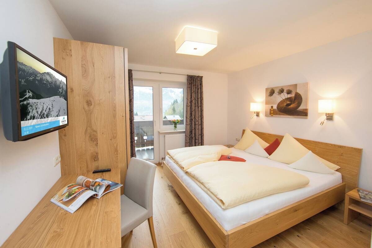 Habitación de hotel con una cama de madera, un escritorio con una revista, un televisor en la pared y un cuadro sobre la cama.