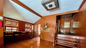 A spacious room with a wooden floor, a bar area with stools, a staircase, and a ceiling light fixture.