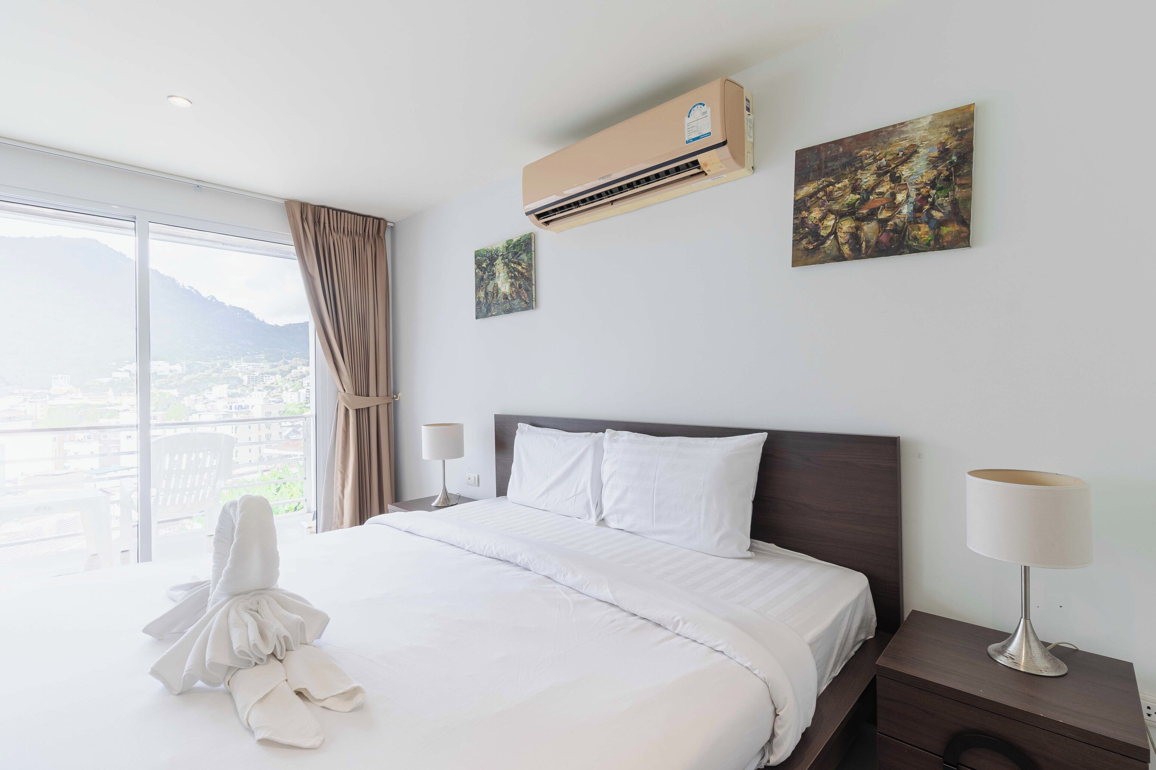 A hotel room with a large bed, an air conditioner, a painting, a lamp, and a view of a cityscape.
