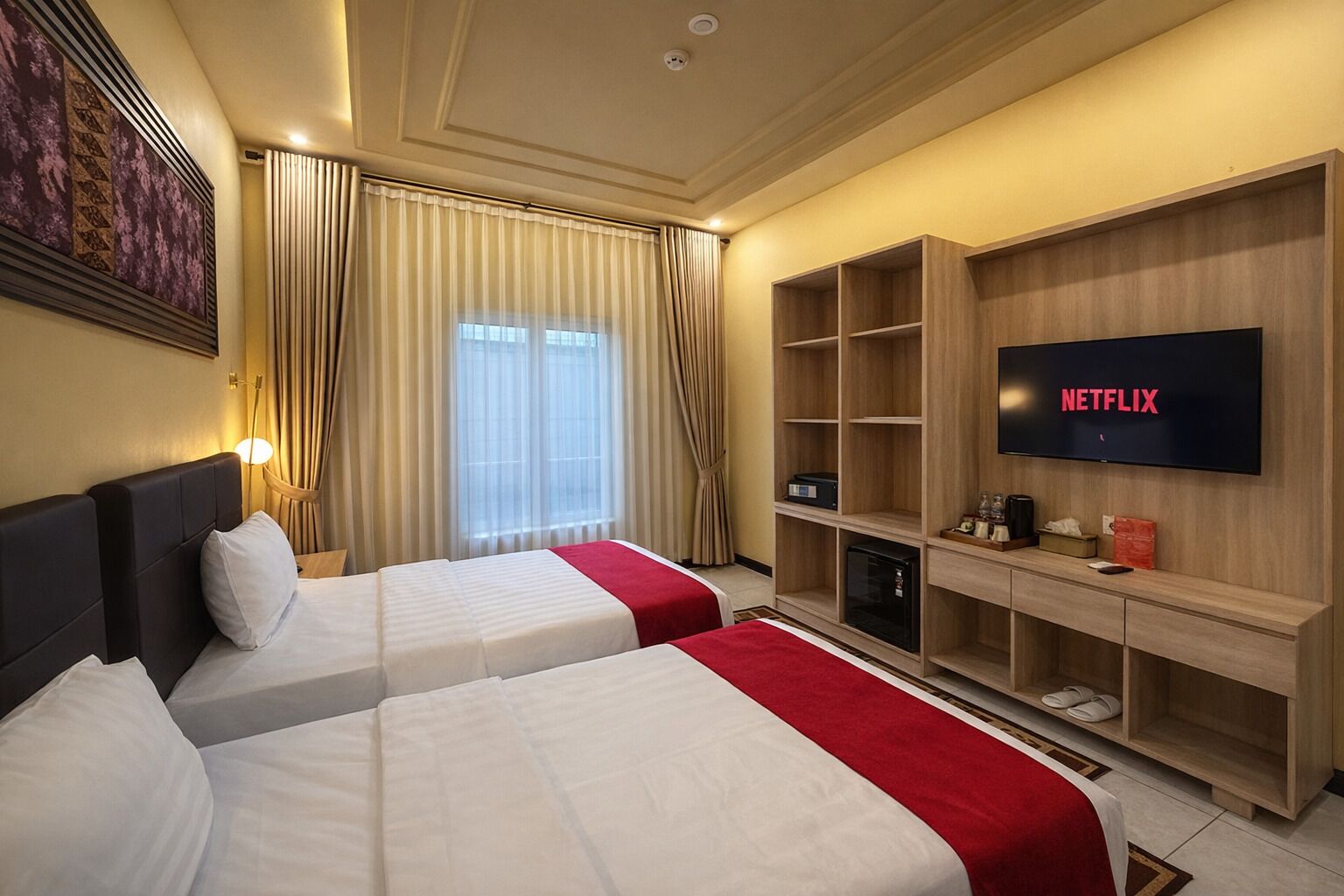 Habitación de hotel con dos camas, televisor, mueble de entretenimiento de madera y ventana con cortinas.
