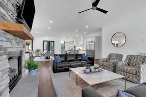 A modern living room with a fireplace, a black leather sofa, a patterned armchair, a marble coffee table, and a kitchen in the background. - Lakefront Farmhouse | 14 Guests | Tims Ford Lake (Winchester)