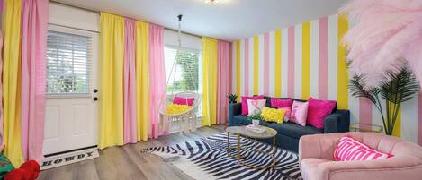 A room with a zebra-patterned rug, a pink armchair, a blue sofa with pink and yellow pillows, and a striped wall.