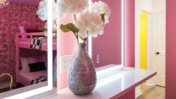 A pink room with a white vase on a white table, a pink bunk bed, and a mirror reflecting the room.