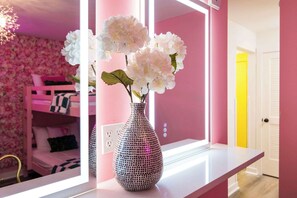 A pink room with a white vase on a white table, a pink bunk bed, and a mirror reflecting the room.