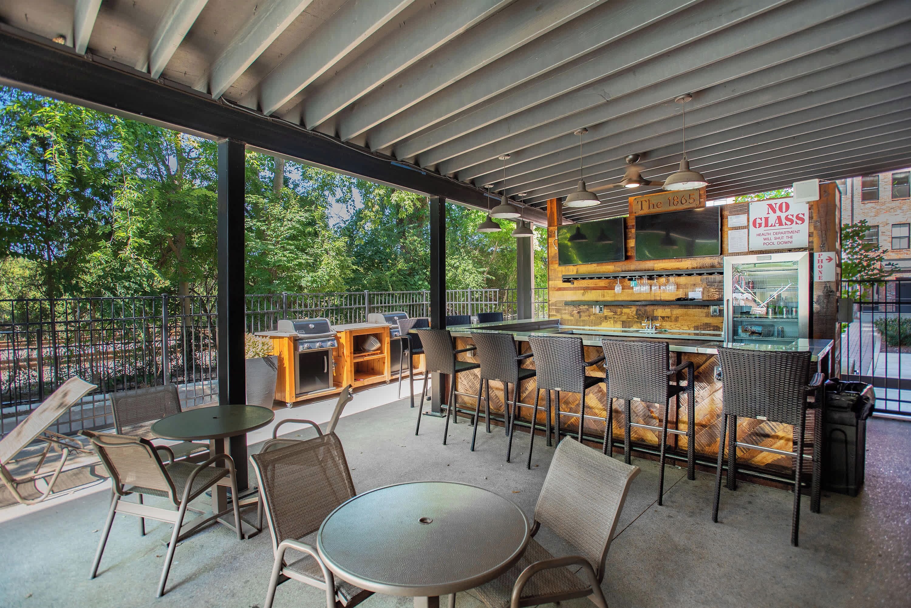 An outdoor patio area with round tables and chairs, a bar counter, and a view of trees.
