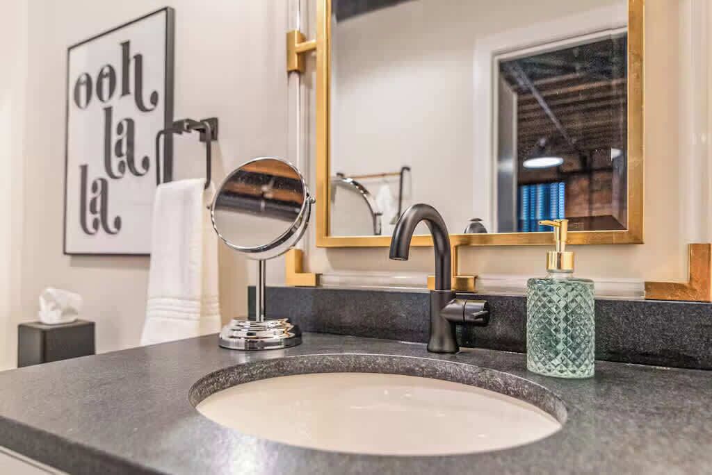 A bathroom with a round mirror, a black faucet, and a green soap dispenser. A framed sign with text is visible on the wall.