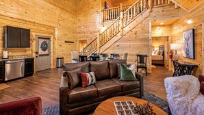 A spacious log cabin interior with a wooden staircase, a leather sofa, a dining area, and a kitchen. - Luxe Cabin • Pool • Hot Tub • Games • Pet Friendly (Pigeon Forge)