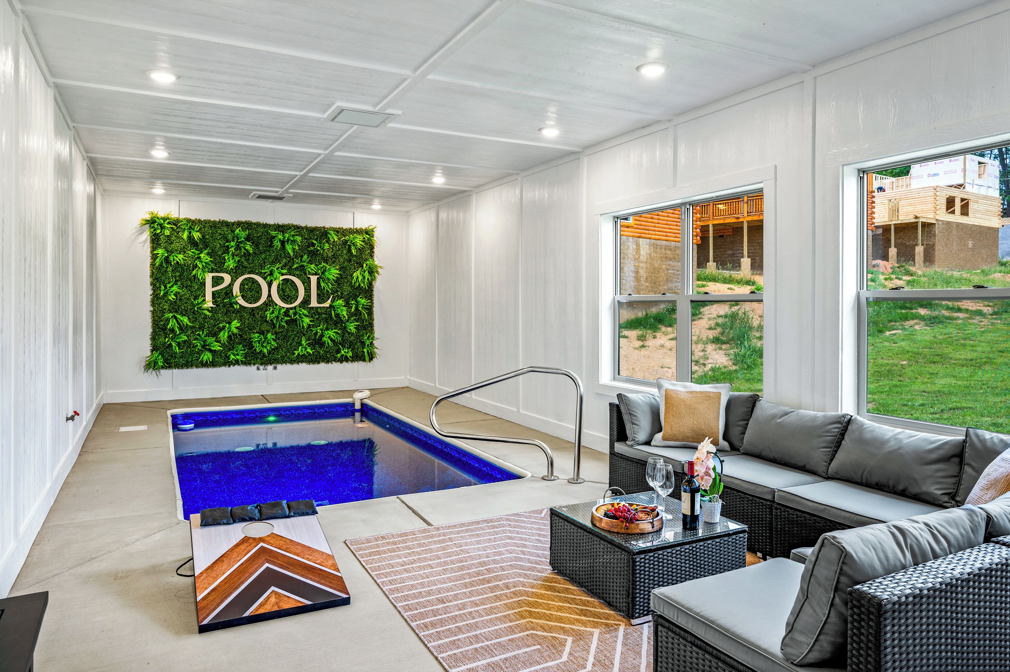 Un espace piscine intérieur moderne, agrémenté d’un mur végétal sur lequel est inscrit le mot « PISCINE », d’un coin salon avec un canapé et une table, et offrant une vue sur l’extérieur grâce à de grandes fenêtres.