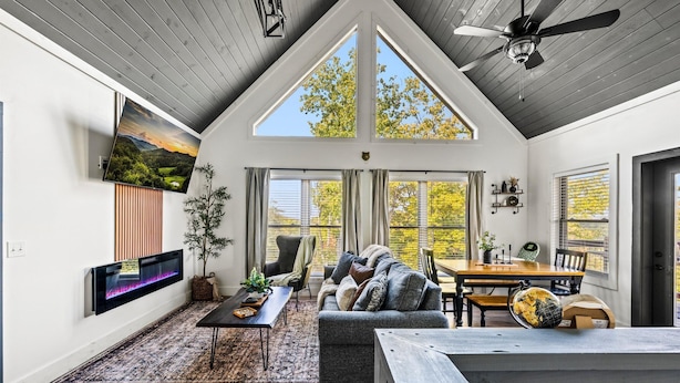 Un vaste salon doté d’un haut plafond, d’un grand téléviseur fixé au mur, d’un coin salon confortable avec un canapé et des fauteuils, d’une table à manger et de grandes fenêtres offrant une vue sur l’extérieur.