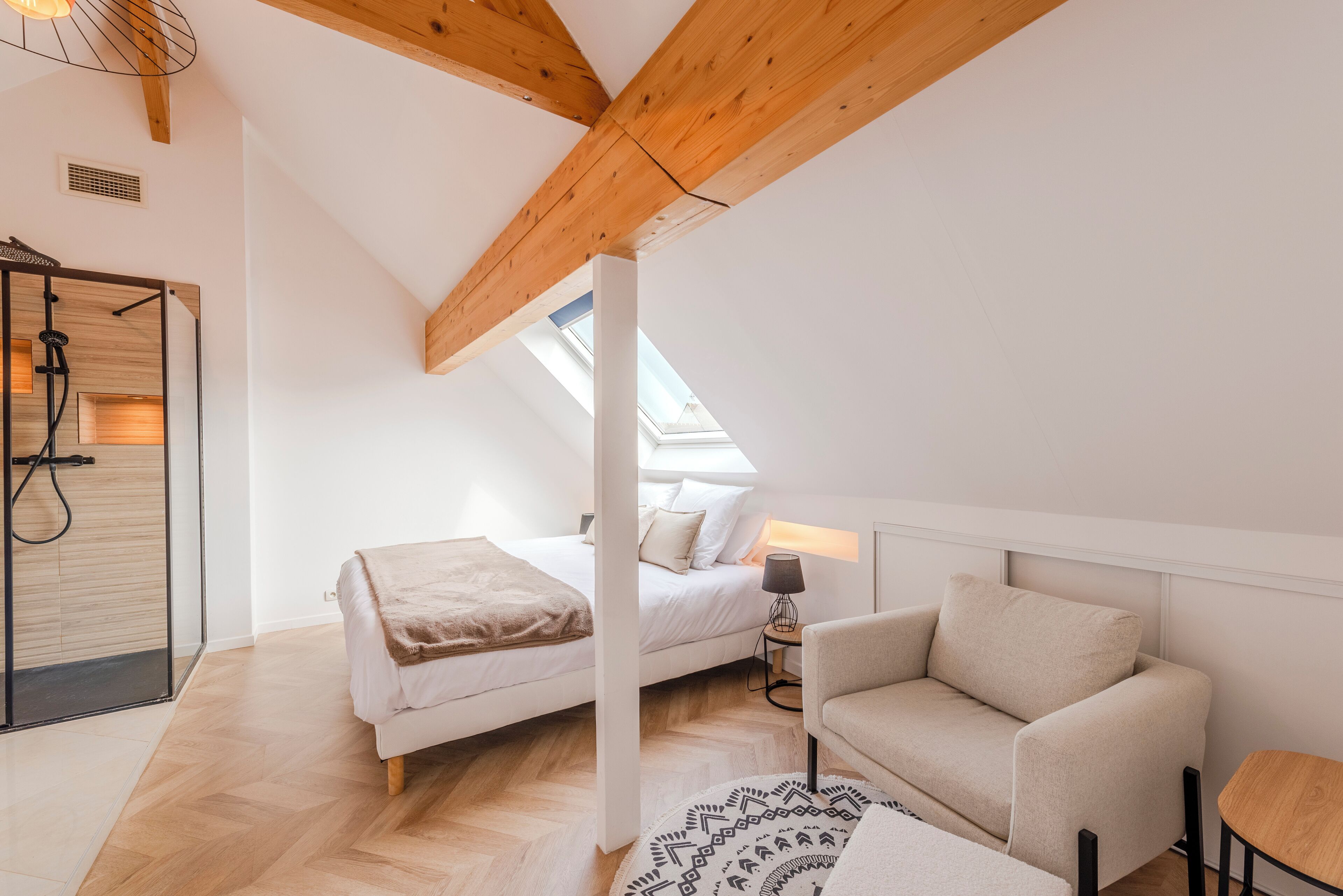 Un dormitorio con cama, una silla, una mesita, ducha y tragaluz.