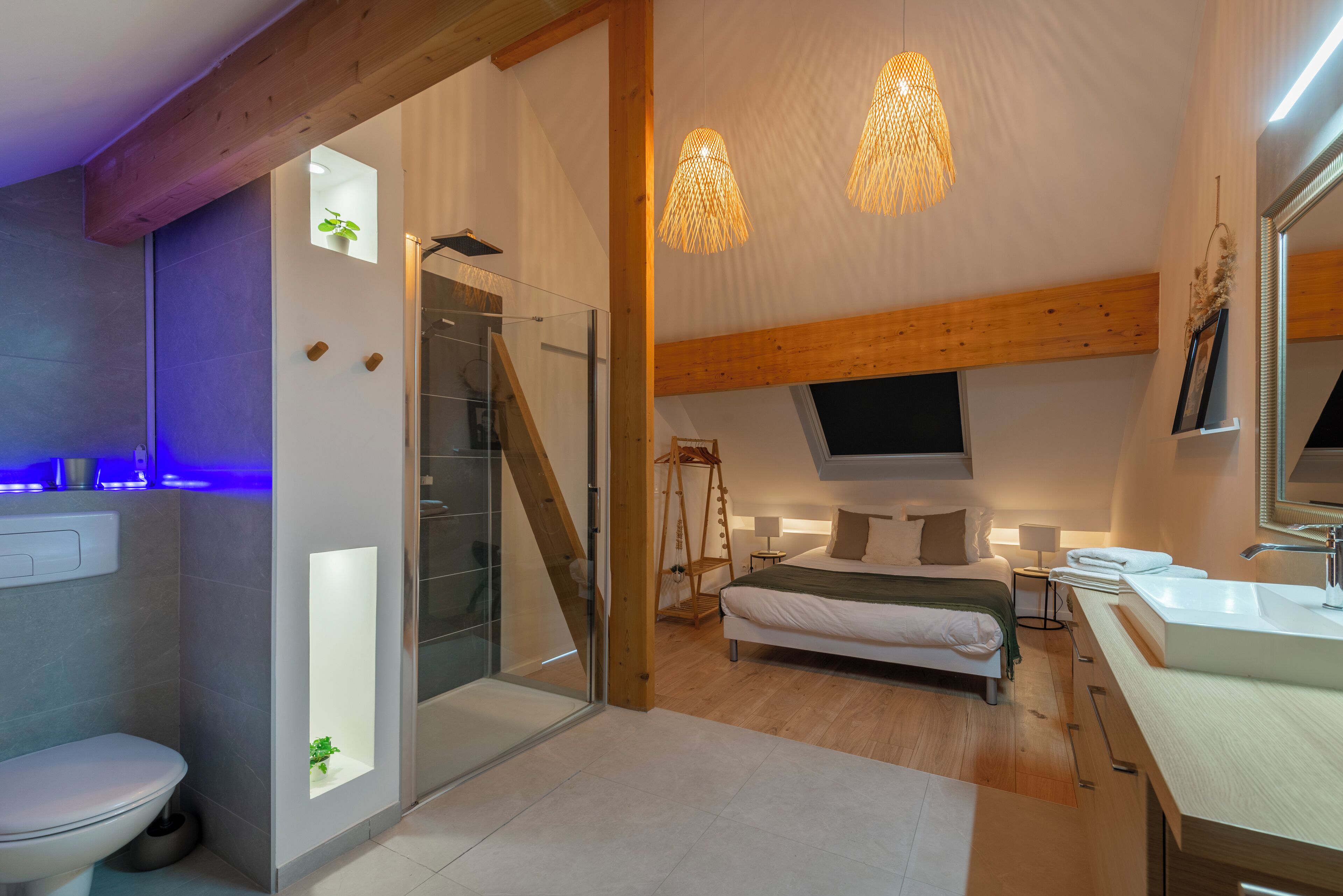 A modern bedroom with a bed, bedside table, and a TV. There is a bathroom with a toilet and a shower. The room has wooden beams and a large window.