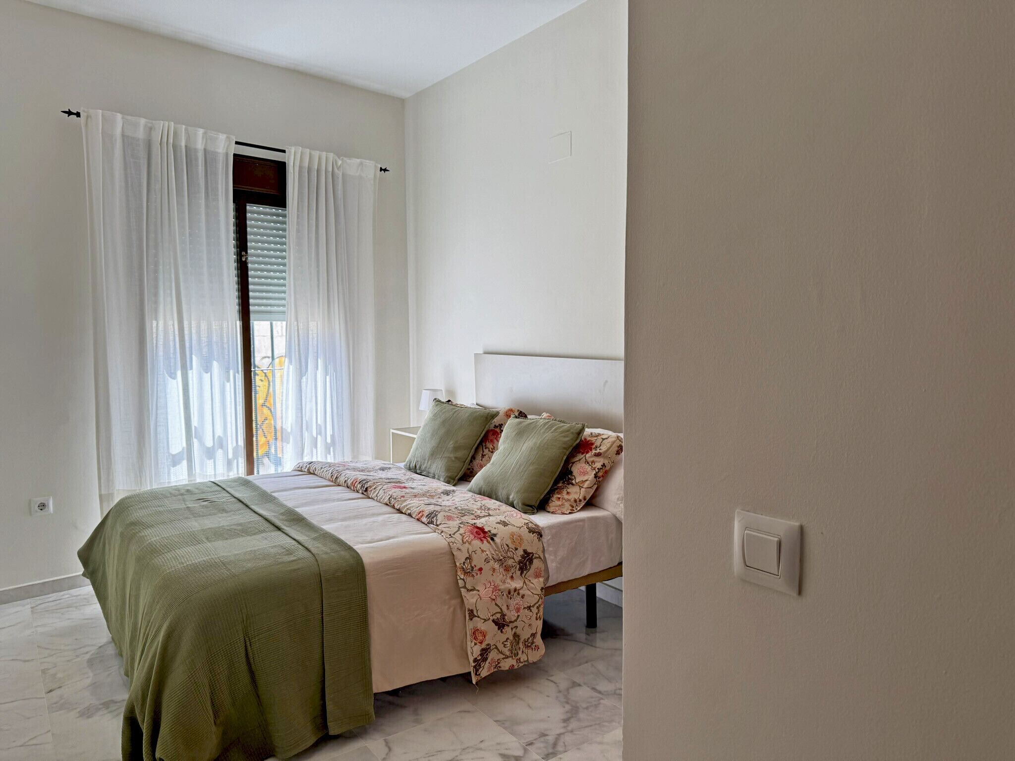 Een slaapkamer met een bed, een raam met gordijnen, een nachtkastje en een lichtschakelaar.