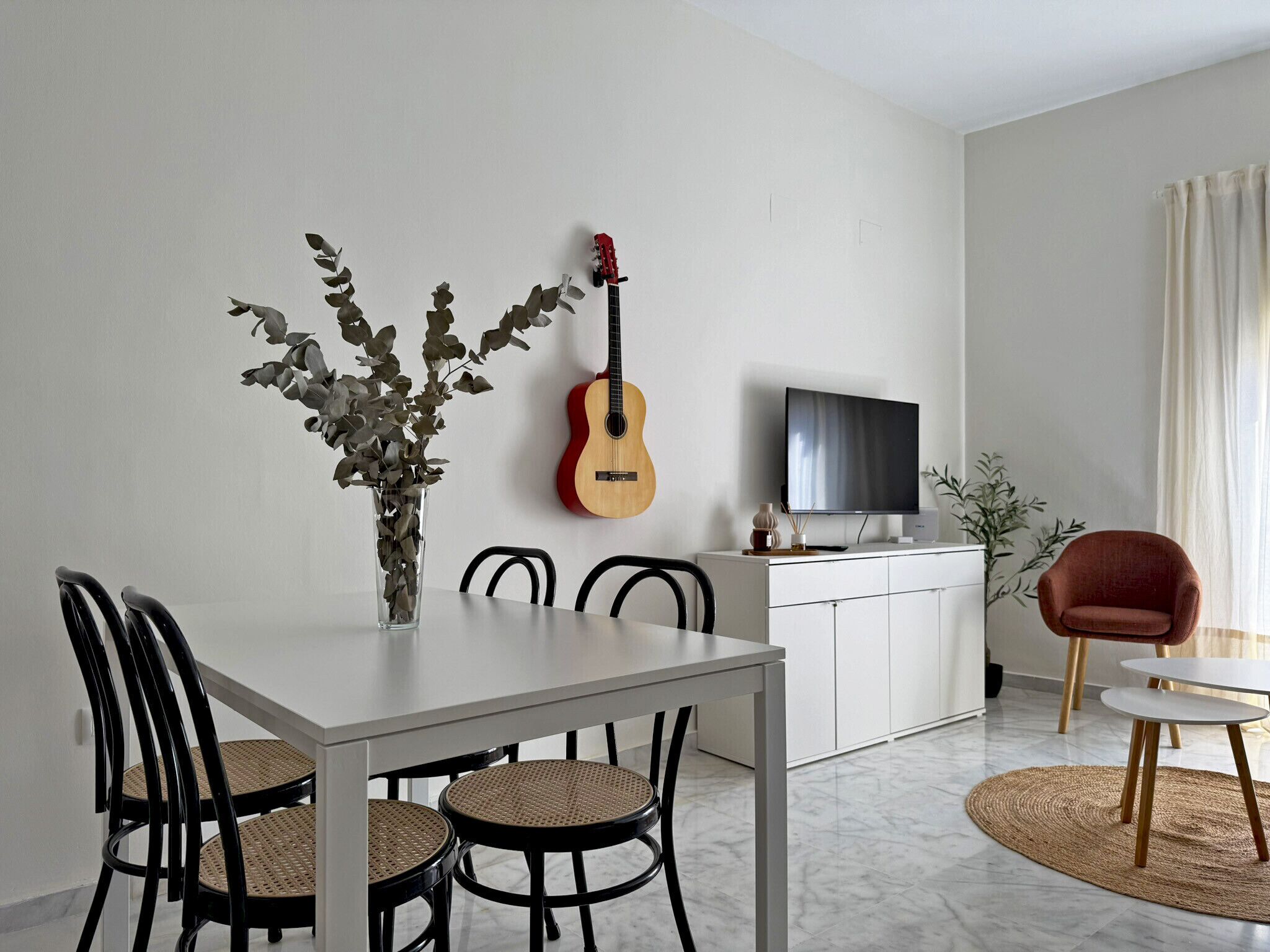 Een eethoek met een witte tafel, zwarte stoelen, een gitaar en een televisie op een kast.