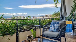 A balcony with a view of the city, a wicker chair with a cushion, a small plant, and a table with a decorative item. - Luxurious Condo at SOHO with Panoramic Romantic Zone Views (Puerto Vallarta)