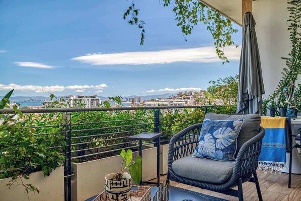 A balcony with a view of the city, a wicker chair with a cushion, a small plant, and a table with a decorative item.