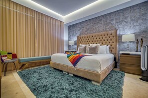 Een moderne slaapkamer met een gestoffeerd hoofdeinde, een groot bed met meerdere kussens, een kleurrijke deken, een blauw, pluizig vloerkleed, een houten bankje, een nachtkastje met een lamp, en een muur met een reliëfpatroon.