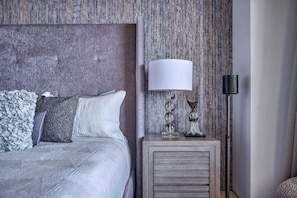 A neatly made bed with a textured white duvet, a wooden headboard, and a bedside table with a lamp and decorative items. - Luxurious Condo at SOHO with Panoramic Romantic Zone Views (Puerto Vallarta)