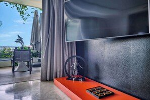 A modern living room with a large flat-screen TV mounted on a textured wall, a red console table with a chess set, a sculpture, and a view of a balcony with outdoor furniture. - Luxurious Condo at SOHO with Panoramic Romantic Zone Views (Puerto Vallarta)
