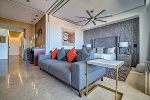Una habitación de hotel moderna con un sofá gris, una mesita de noche, un ventilador de techo y una cama grande con cabecero gris.